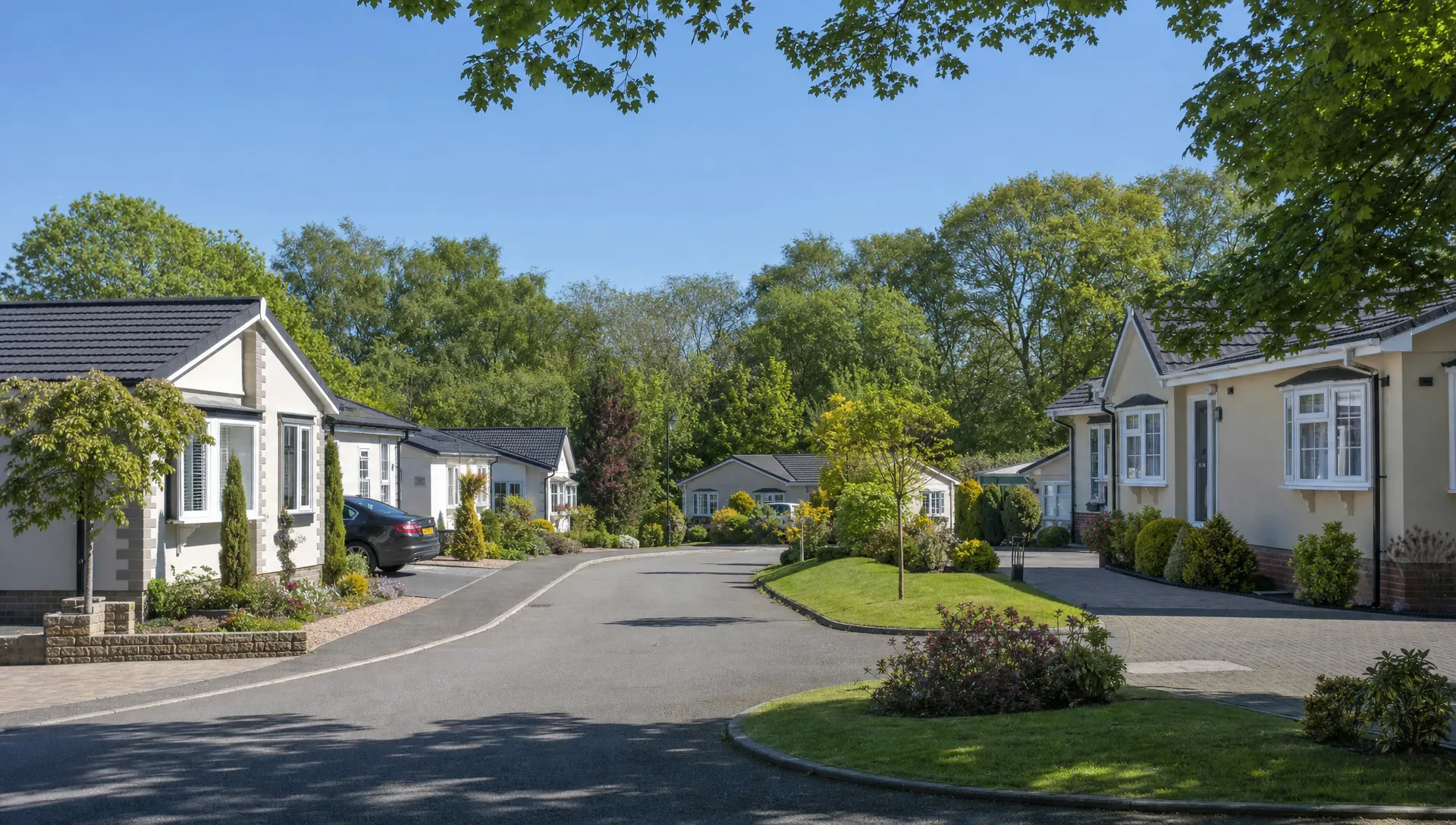 A quiet landscaped lane at Hermitage Park with single-storey luxury park home bungalows, private driveways, neat gardens and mature trees.