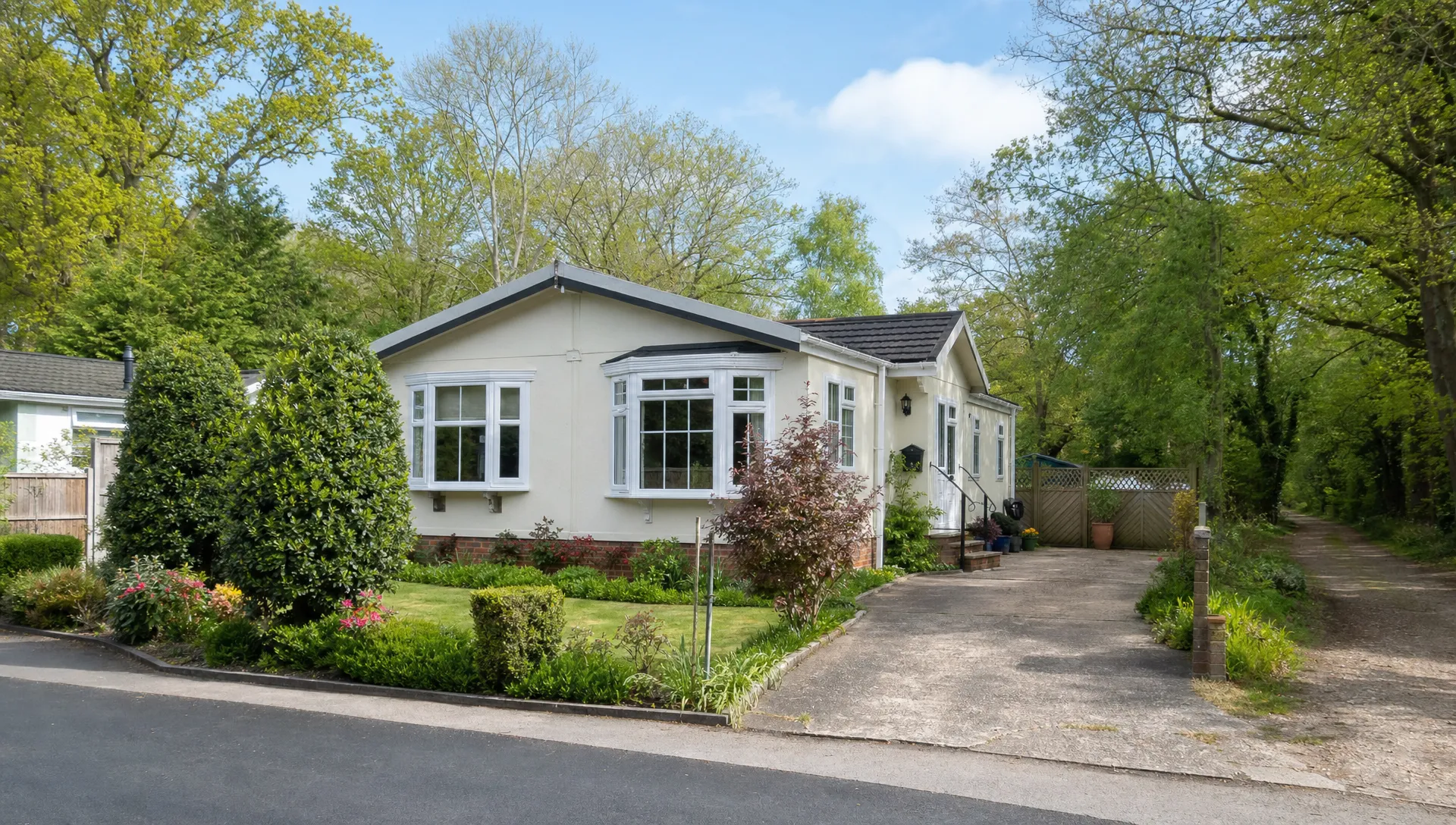 A single-storey park home bungalow in Berkshire with a private garden, driveway and mature trees, seen from the front on a quiet residential plot with a woodland path nearby.